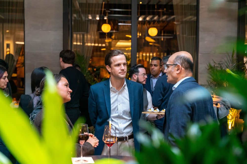 Professionals networking at an upscale indoor cocktail party, engaged in conversation among lush greenery.