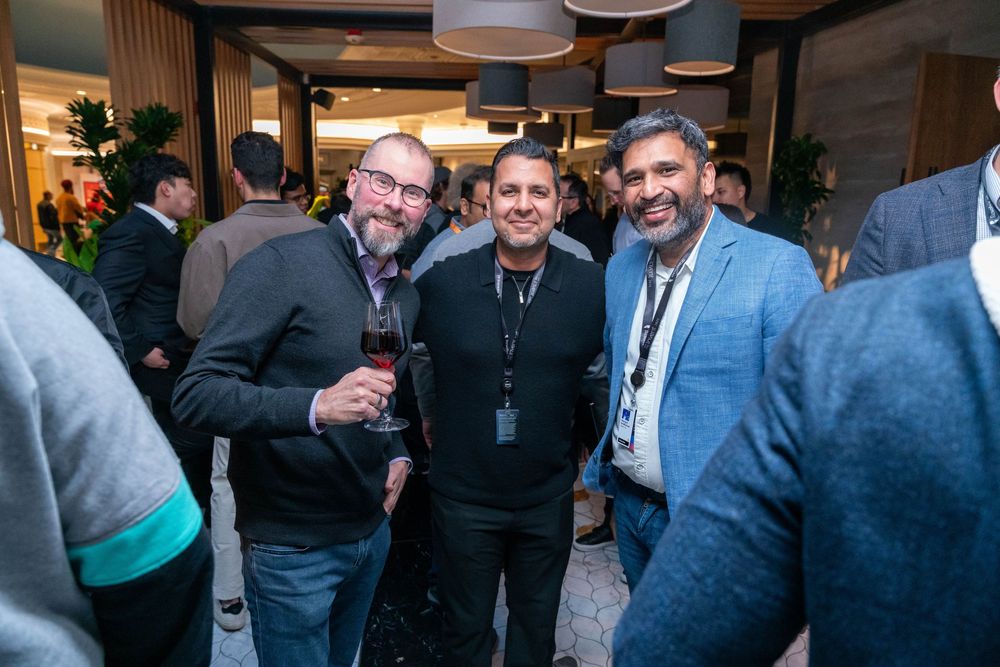 Three men smiling and posing together at a corporate networking event, one holding a glass of red wine.
