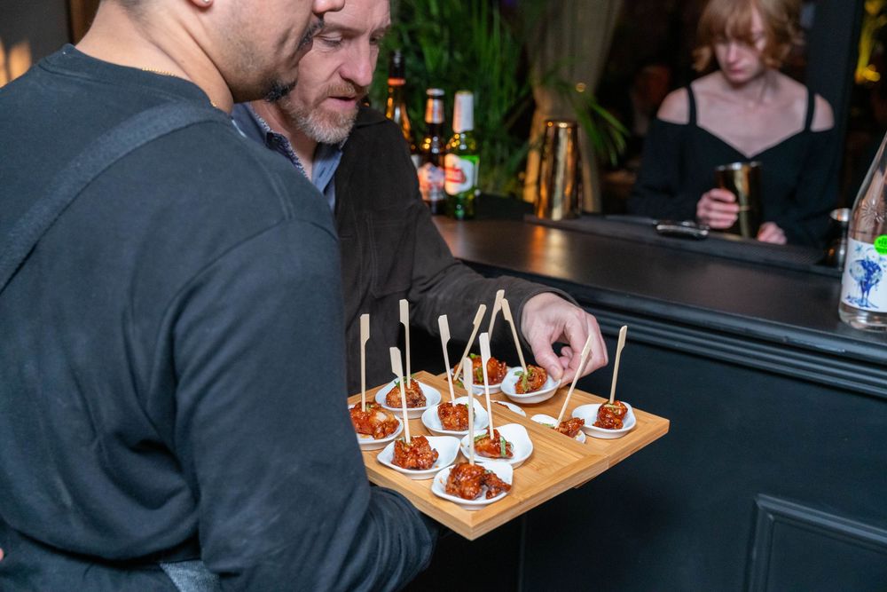 Two men selecting appetizers from a wooden tray of skewered bites at an upscale bar event.