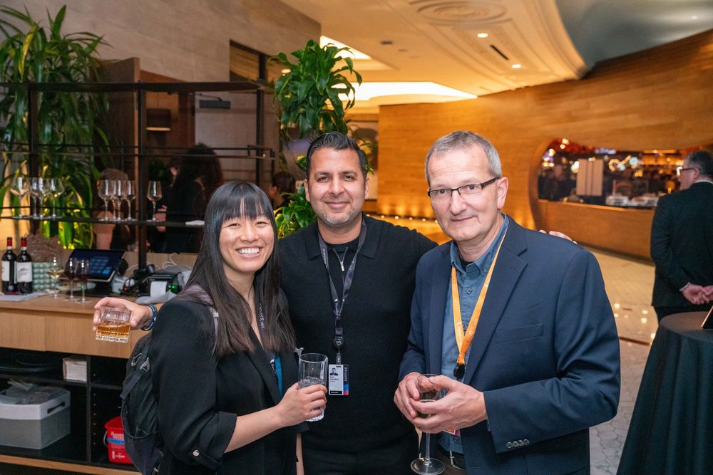 Three professionals smile at a corporate networking event, wearing lanyards and holding drinks in an elegant venue.
