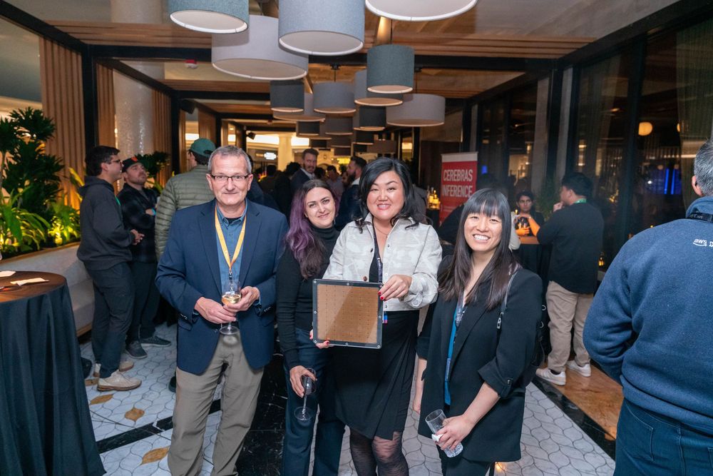 Four people posing together at an indoor awards event, one holding a plaque, with guests mingling in the background.