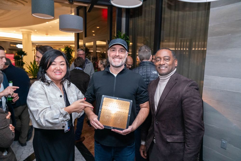 Three professionals smiling and holding a wooden award plaque at a corporate recognition event indoors.