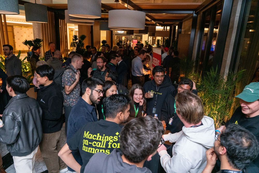 Crowd of diverse attendees networking at a tech event in a modern venue, one wearing a Machines Call for Humans shirt.
