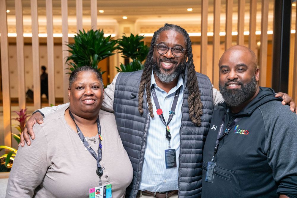 Three smiling professionals wearing lanyards pose together at an indoor corporate event or conference.