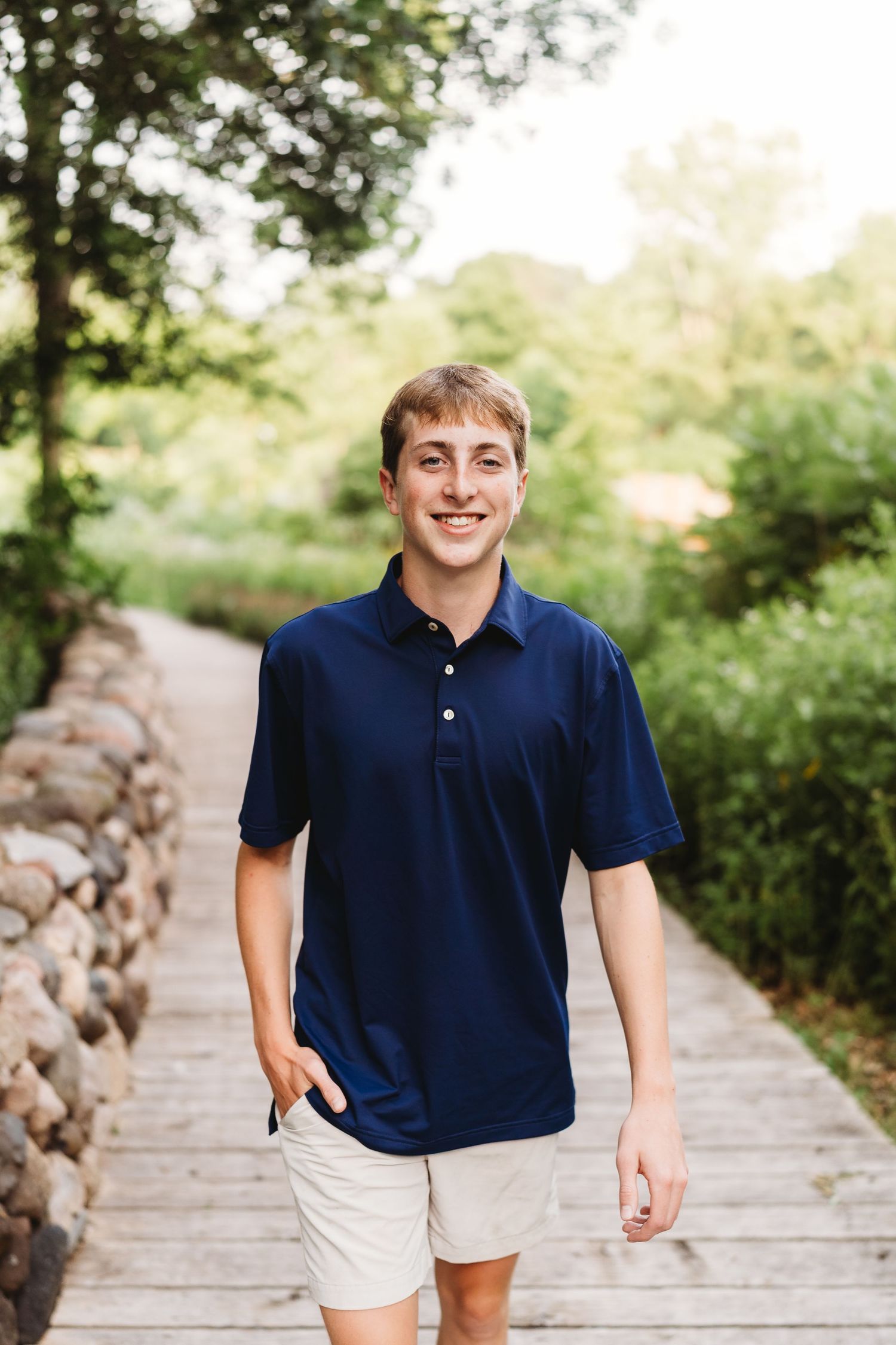 Young man in navy polo shirt and khaki shorts smiling while walking on a wooden path surrounded by greenery.
