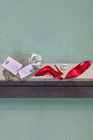 Red high heels and wedding stationery with tropical leaf motifs displayed on a ledge by a green tiled pool.