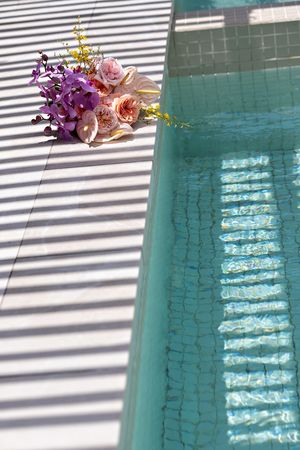 Floral bouquet with pink roses and purple orchids resting beside a sunlit turquoise swimming pool.