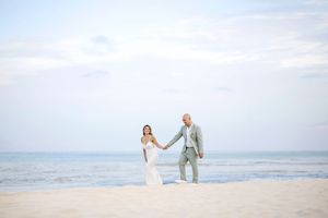 Bride and groom holding hands on a sandy beach with ocean waves and soft blue sky in the background.