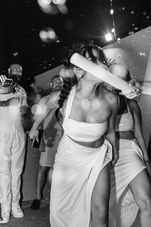Black and white photo of a woman in white crop top and skirt dancing at a party with bubbles.