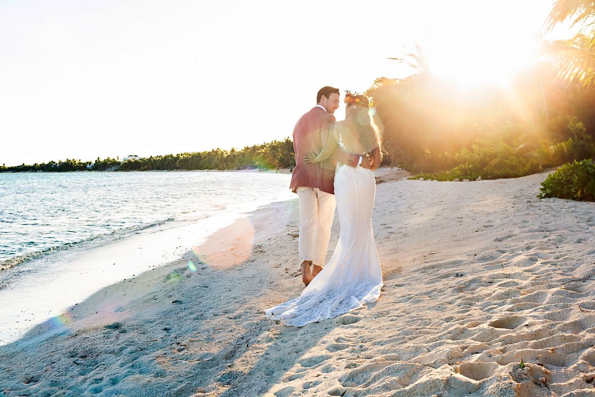 Bride and groom embrace on a sunny tropical beach at sunset, with golden light and turquoise water in the background.