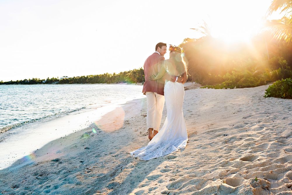 Bride and groom embrace on a sunny tropical beach at sunset, with golden light and turquoise water in the background.