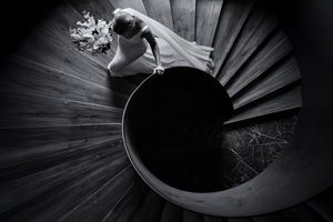 Black and white aerial view of a bride in a white gown descending a spiral staircase, holding a bouquet.