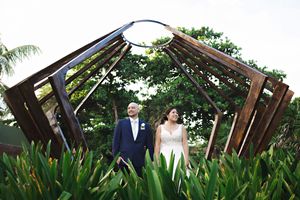 Bride and groom stand beneath a rustic wooden arch sculpture surrounded by lush tropical greenery at an outdoor wedding.