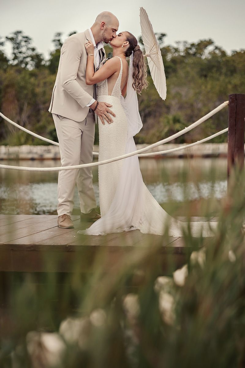 Bride and groom share a romantic kiss on a wooden dock surrounded by water and lush greenery on their wedding day.