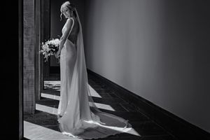 Bride in elegant white gown and veil holding bouquet, standing in dramatic doorway light, black and white photo.