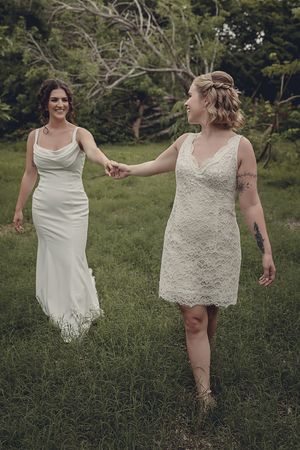 Two brides in white wedding dresses hold hands while walking through a lush green outdoor setting.