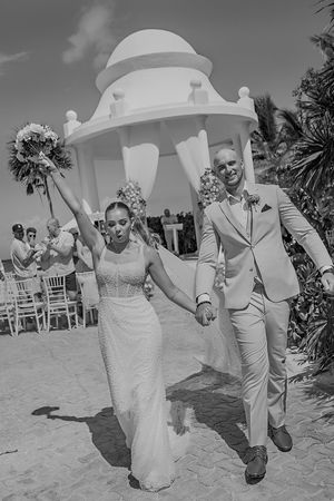 Black and white photo of a joyful newlywed couple holding hands, walking away from a beach wedding gazebo.
