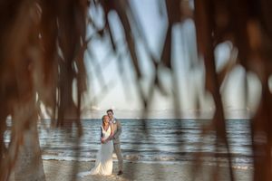 Bride and groom embrace on a beach at sunset, framed by tropical palm fronds with ocean waves behind them.
