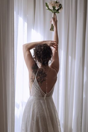 Bride in backless lace wedding dress holding bouquet aloft near sheer curtains, showing elegant back tattoo.