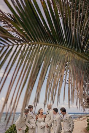 Bride and groomsmen in tan suits laughing on a tropical beach, framed by a lush palm frond with ocean views.