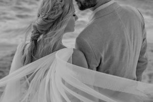 Black and white photo of a bride and groom embracing by the ocean, her veil flowing in the wind.