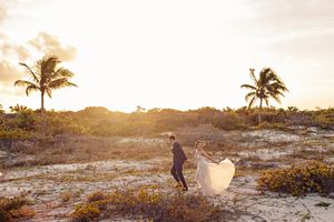 Bride and groom walk on sandy beach at golden sunset with tropical palm trees in romantic destination wedding photo.