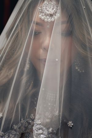 A bride wearing a pearl headpiece and embroidered veil, her face partially visible beneath the sheer white fabric.