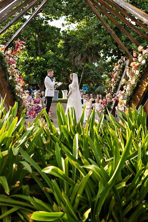 Outdoor wedding ceremony under floral arch with bride and groom exchanging vows surrounded by lush greenery.
