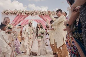 Indian wedding couple exits ceremony under floral arch on beach, guests celebrating with flower petals.