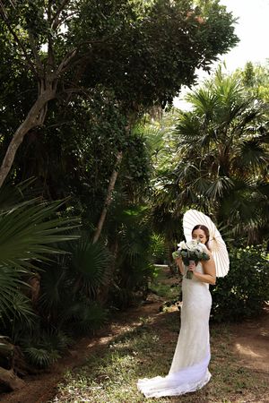 Bride in white lace gown holding bouquet and parasol umbrella, standing in lush tropical garden setting.