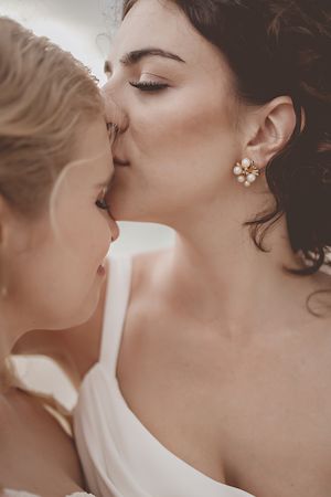 Two brides share a tender kiss, one wearing pearl flower earrings, in an intimate wedding moment.