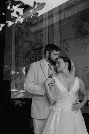 Black and white wedding photo of bride and groom embracing tenderly near a glass window with map reflection.