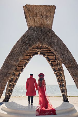 Couple in red attire holding hands under thatched arch structure by the ocean beach.