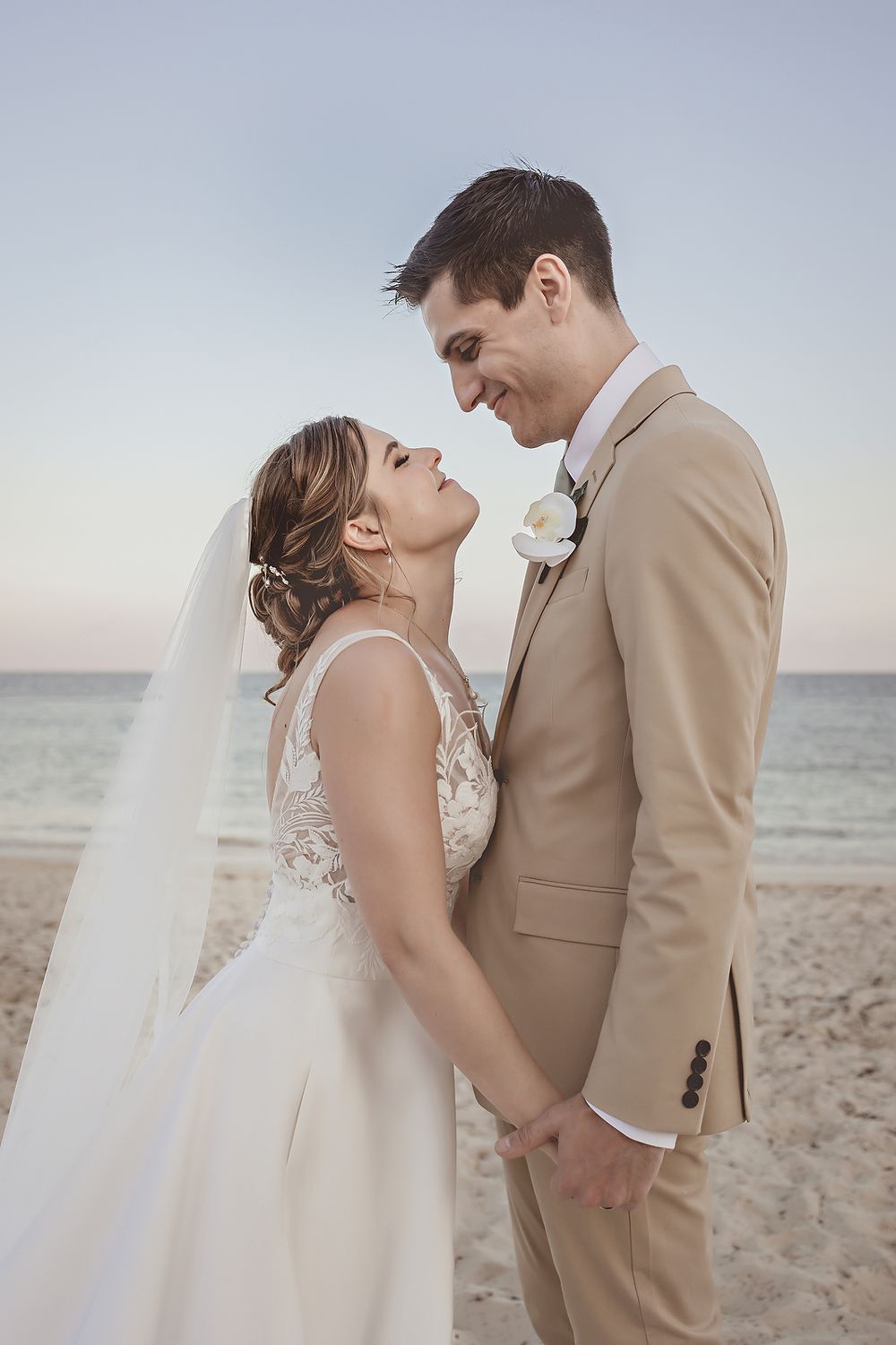 Bride and groom holding hands on a sandy beach at sunset, smiling at each other in wedding attire.
