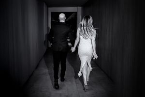 Black and white photo of a couple walking hand in hand down a hallway, woman in white dress, man in dark suit.