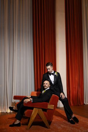 Two grooms in black tuxedos pose elegantly by red velvet curtains, one seated in a velvet chair.