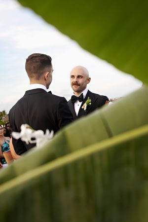 Two grooms in black tuxedos share a heartfelt moment during their outdoor wedding ceremony framed by tropical leaves.
