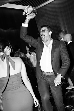 A man in a suit dances energetically at a party, raising a shoe overhead with his tongue out, in black and white.