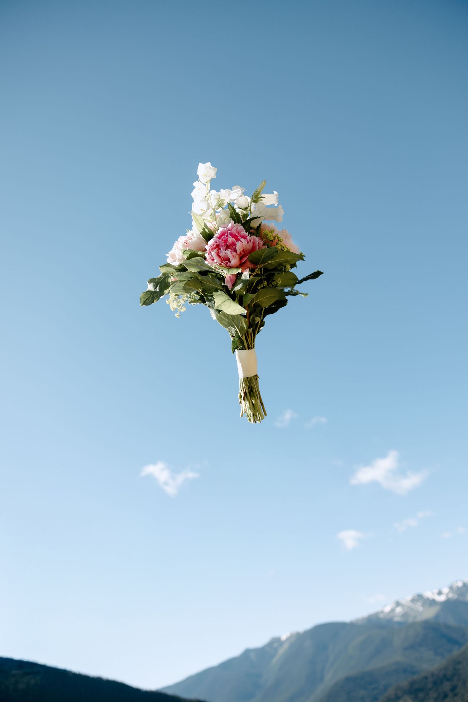 A bridal bouquet with pink peonies and white flowers tossed against a clear blue sky with mountains.