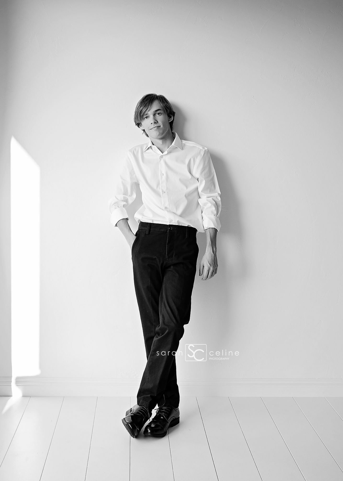 Black and white portrait of a young man in white shirt and black pants leaning against a white wall.