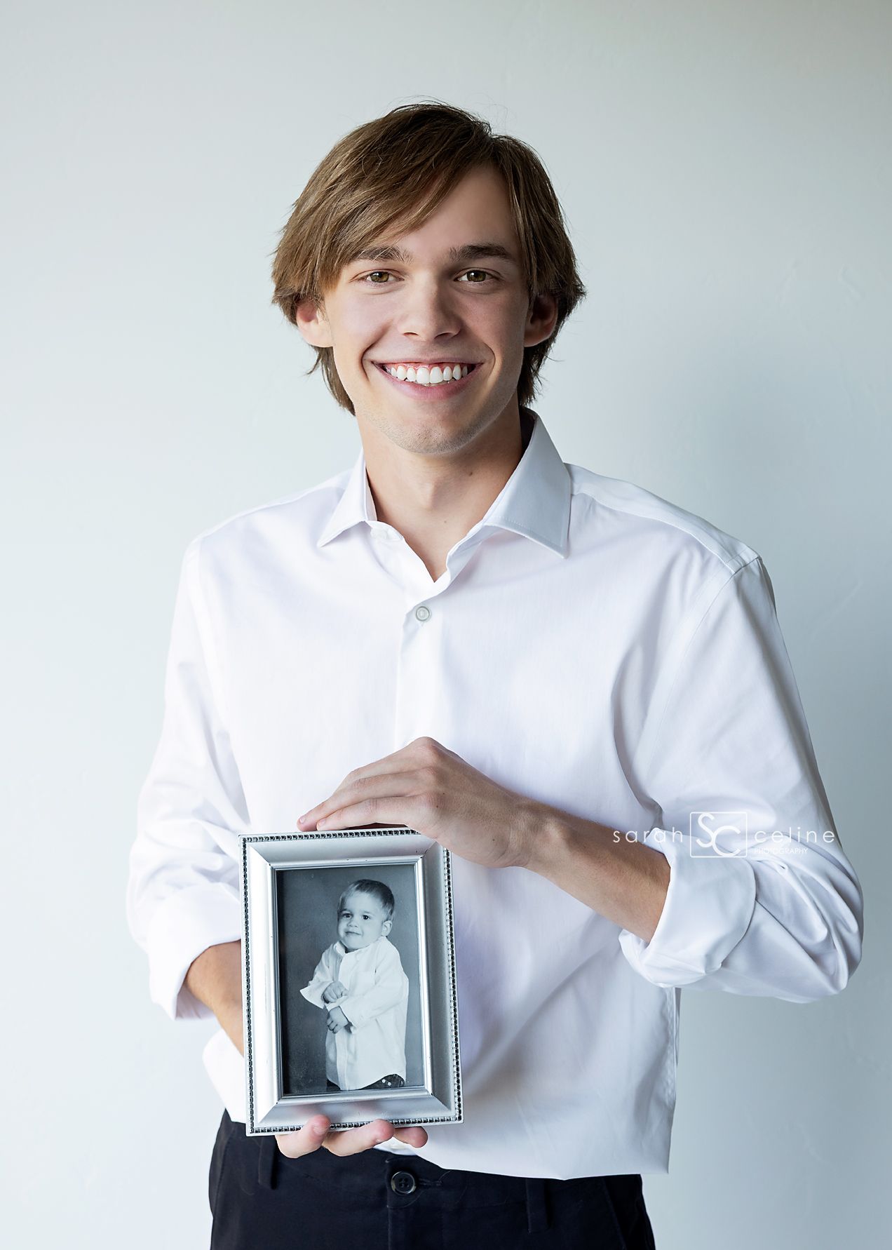 Smiling teenage boy in white shirt holding a framed black and white baby photo against a light background.