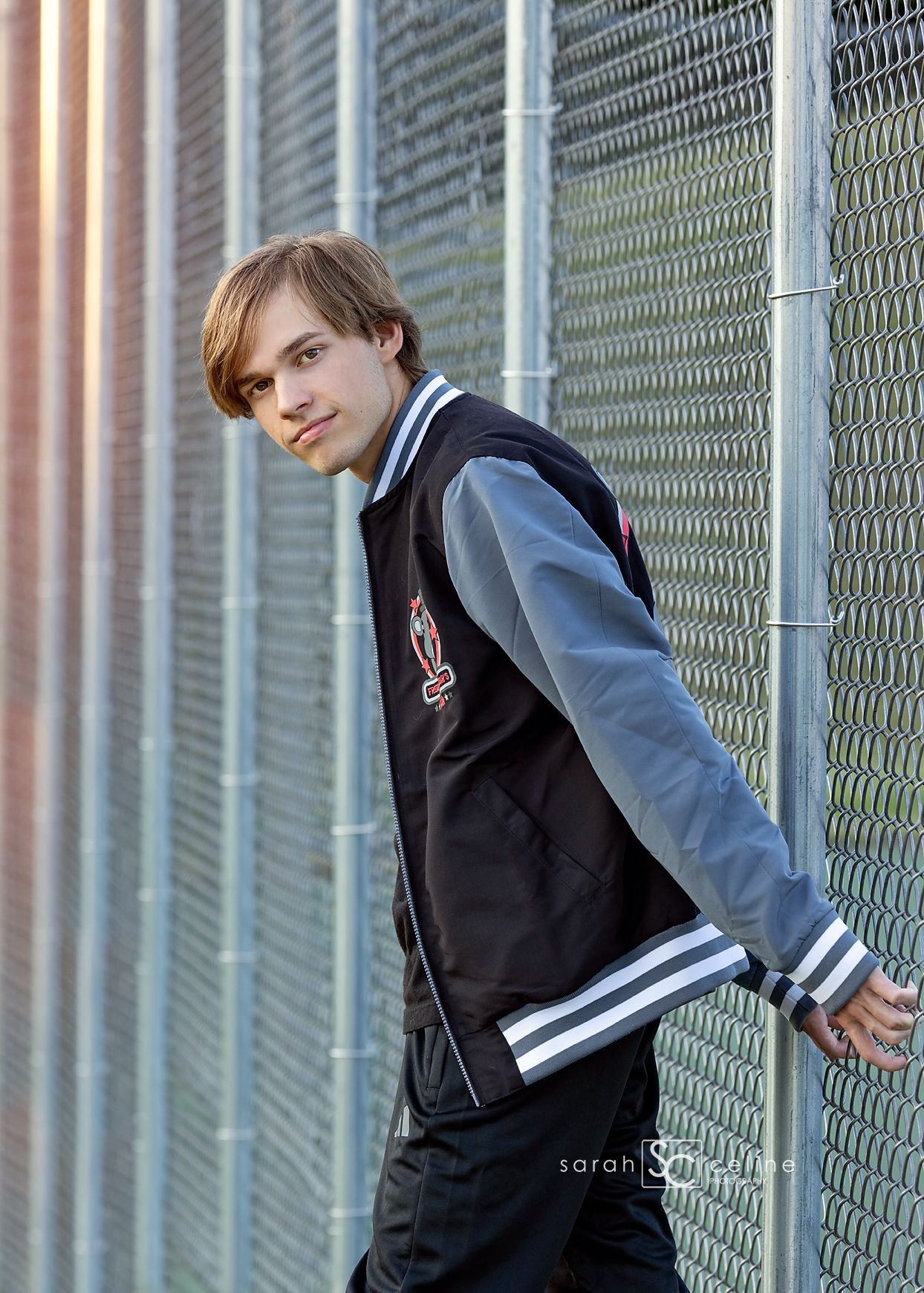 Young man in black and gray varsity jacket leaning against chain-link fence in professional senior portrait photo.