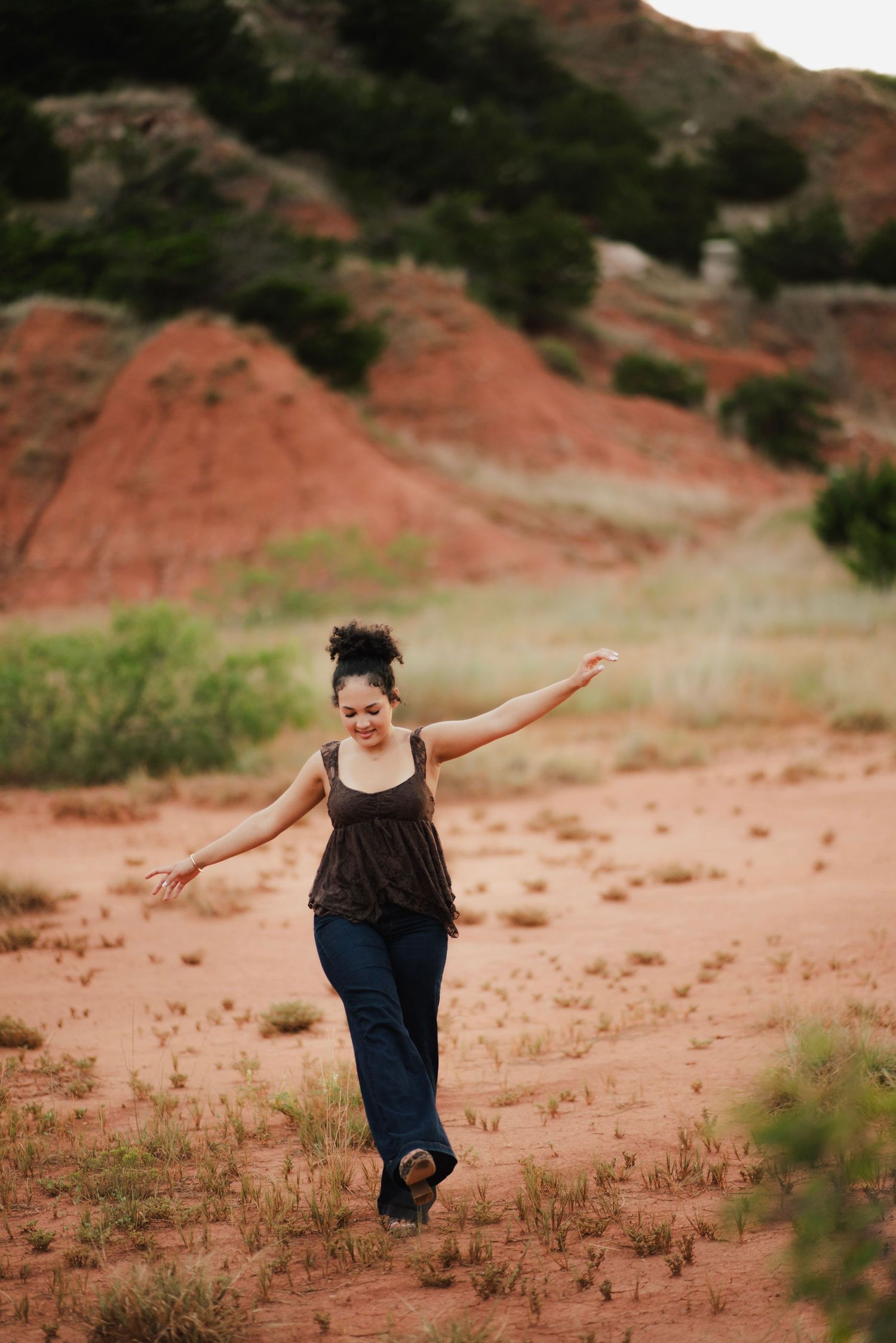 Woman in jeans and tank top dancing freely on red desert sand with scenic red rock formations behind her.
