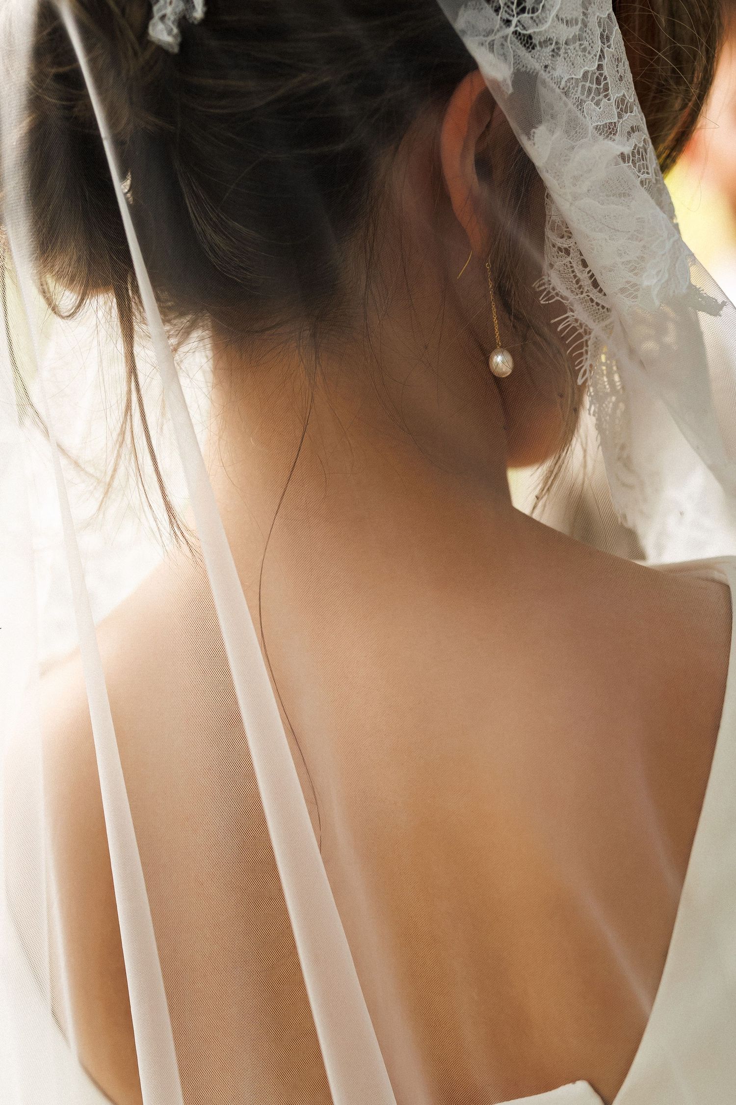 Bride wearing a white lace veil and pearl drop earring, photographed from behind showing her bare back and updo hairstyle.