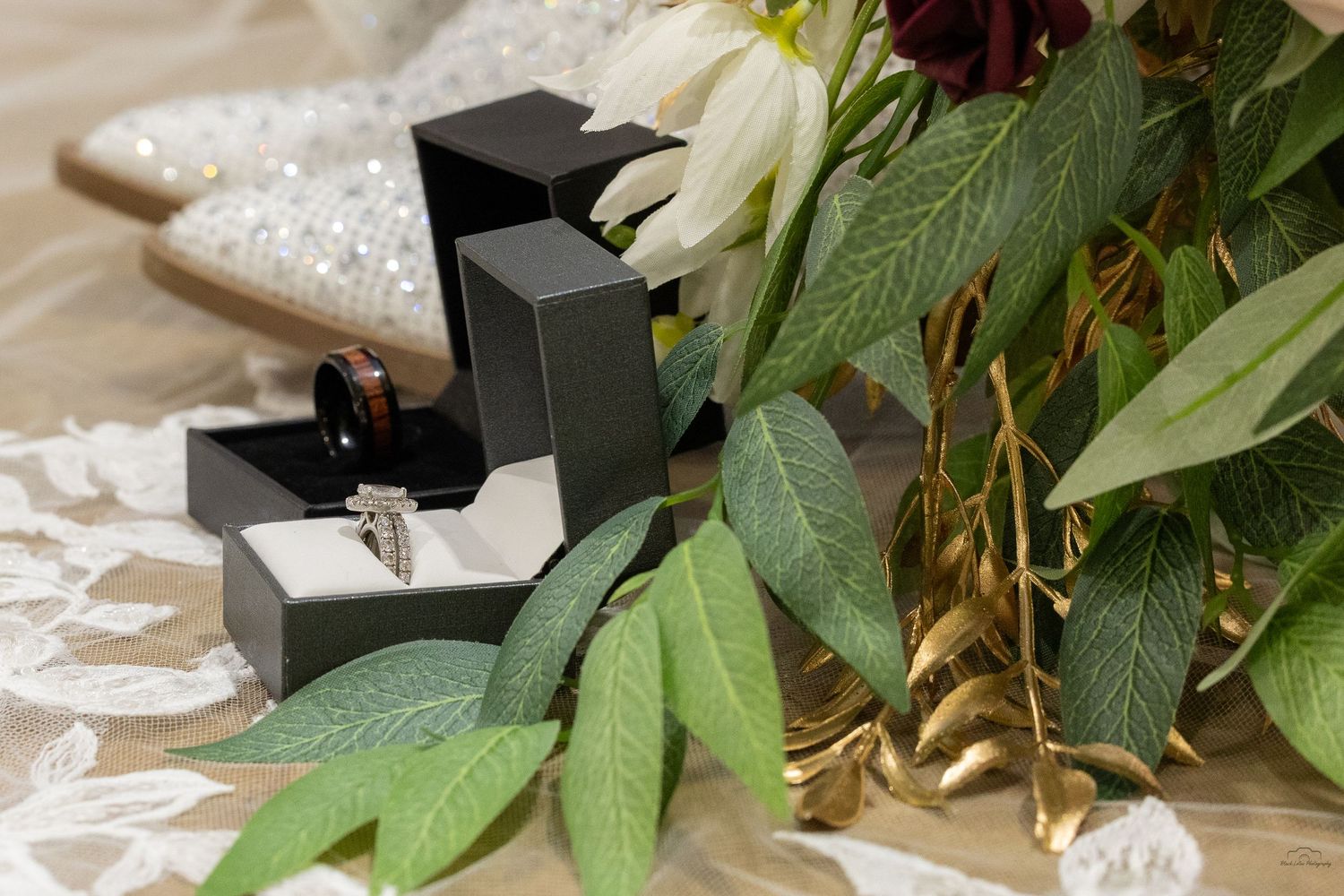 Diamond ring in black box beside bridal bouquet with red and white flowers, green leaves, and white lace veil.