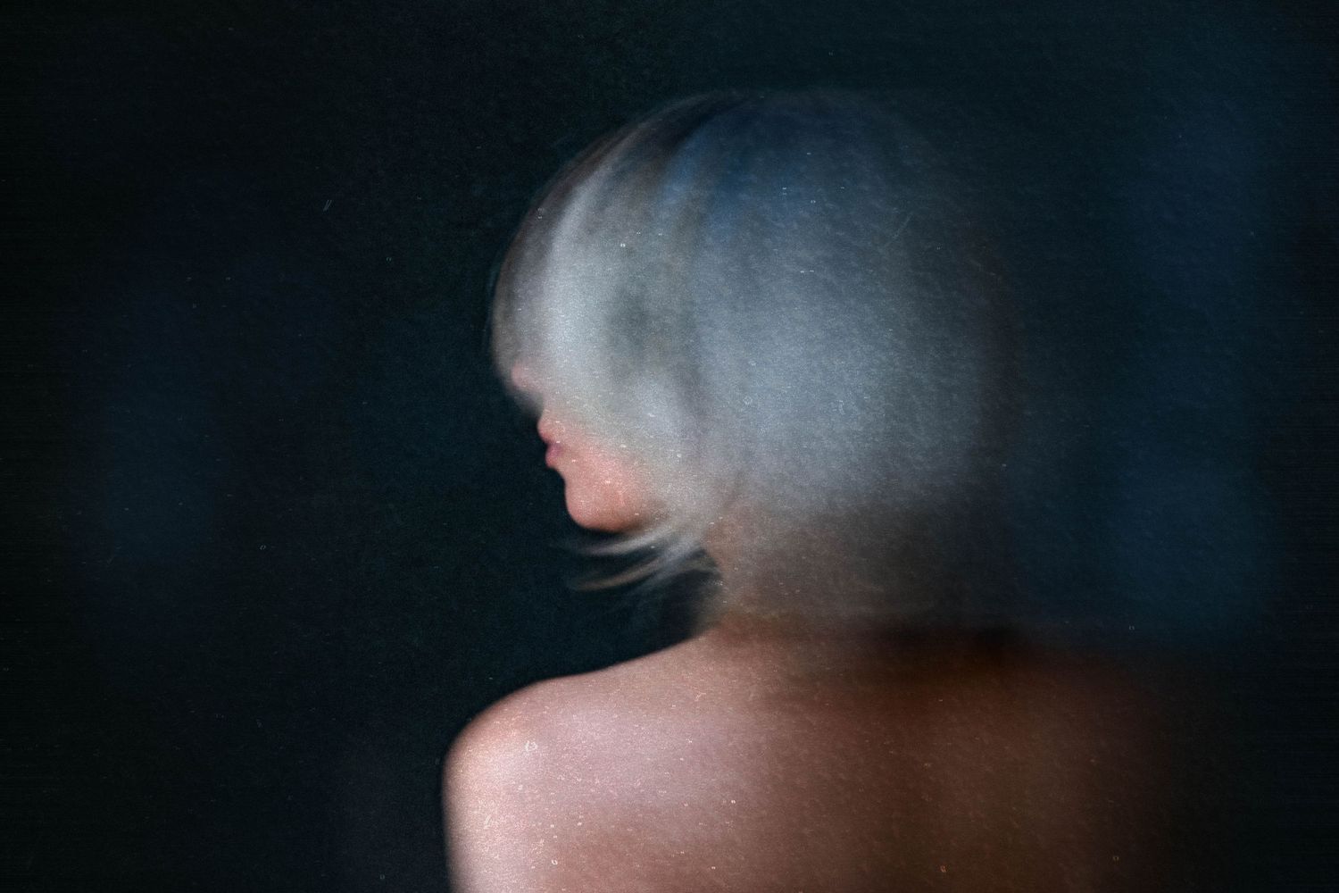 Blurred motion portrait of a person with short silver hair against a dark background.