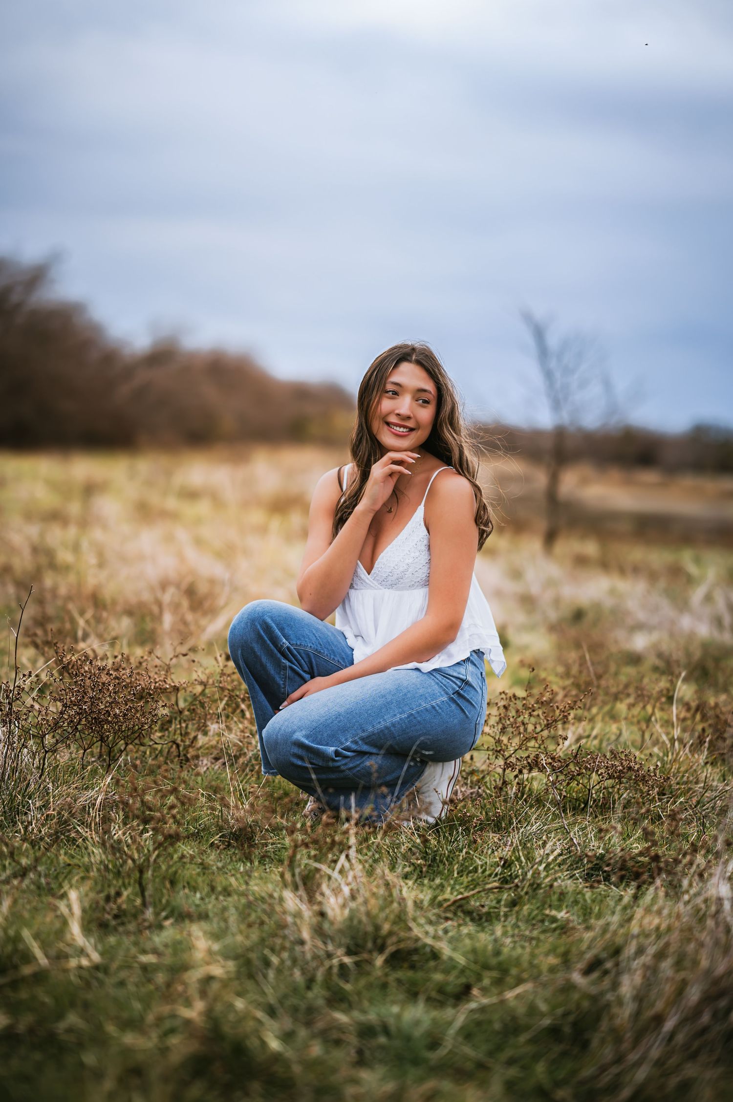 A teenage girl in a white top and jeans crouches in an open grassy field under a cloudy sky.