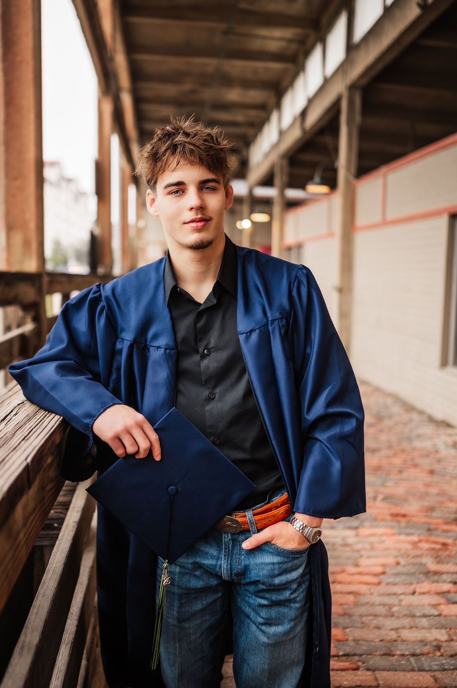 A young man in a blue graduation gown and cap leans on a railing in an urban outdoor setting.