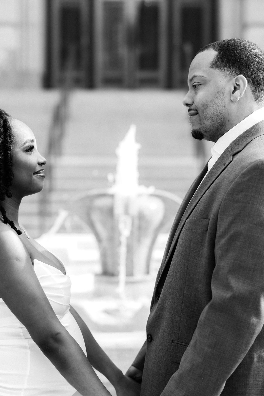 Black and white photo of a couple holding hands and gazing at each other in front of a fountain.