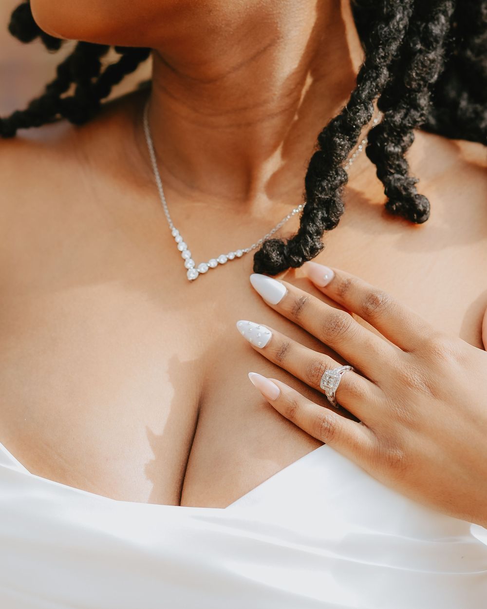 Close-up of woman with braids wearing a diamond V-necklace, ring, and white manicured nails on chest.